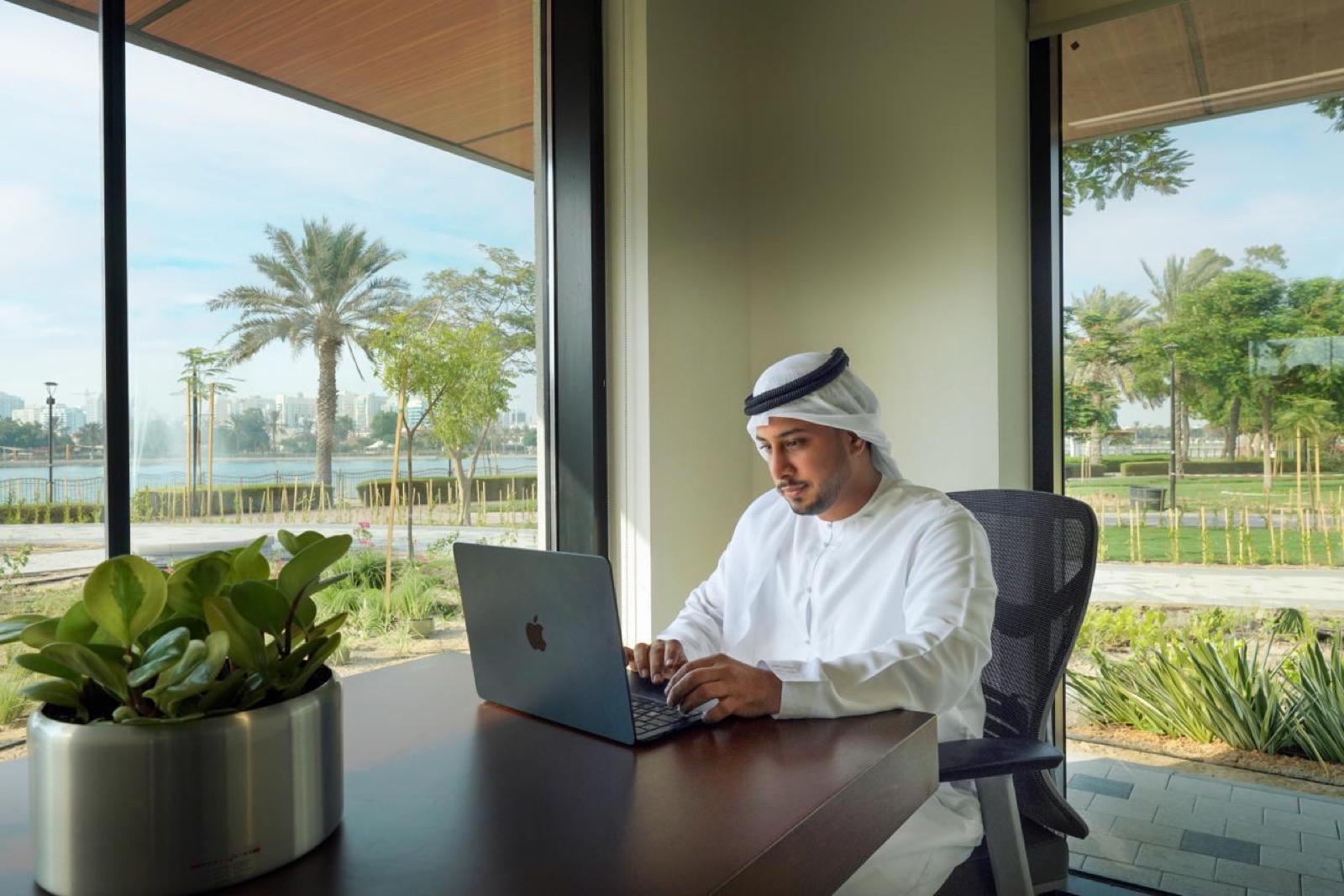 A member in kandura working on a laptop by a floor-to-ceiling window overlooking a green park.