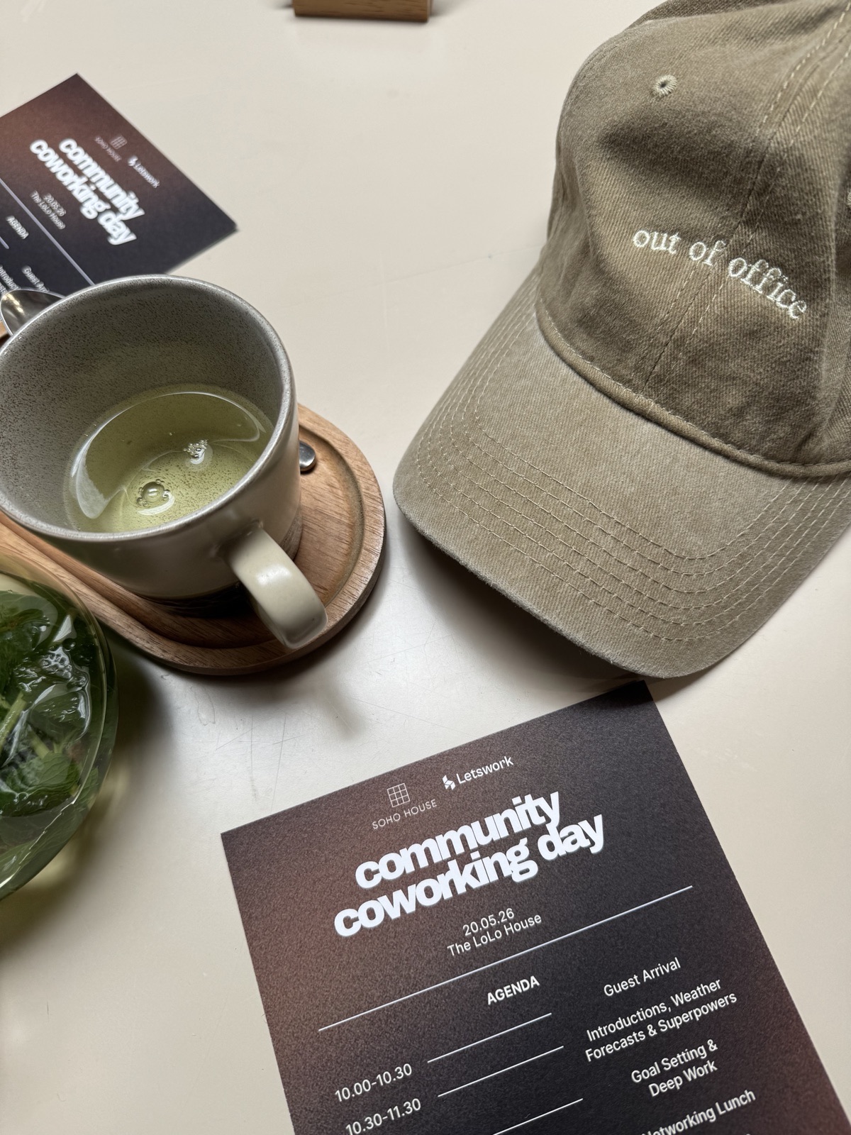 Flatlay of a Soho House × Letswork 'community coworking day' agenda card, an 'out of office' cap and a matcha.